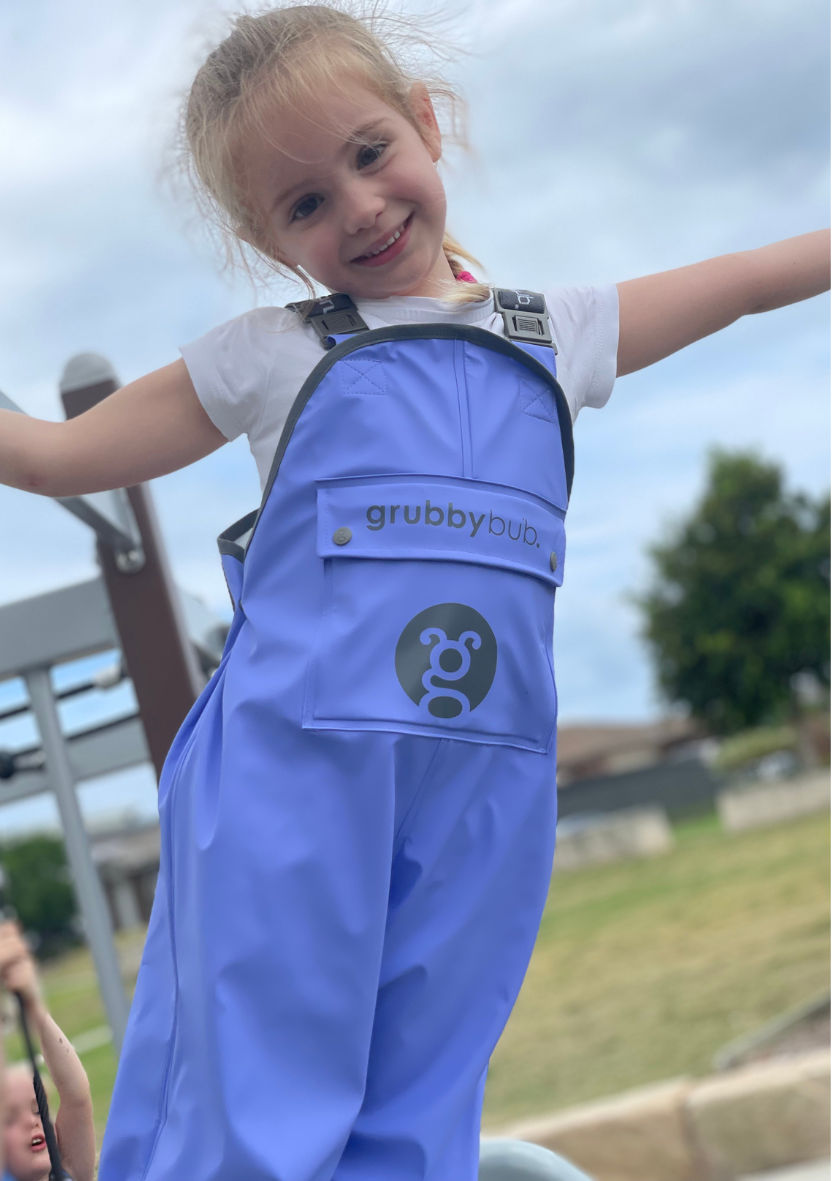 toddler girl in waterproof rain pants hanging sideways from playground equipment